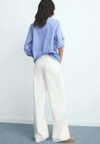 Vrouw met lang donker haar, gekleed in een blauw gestreept shirt en wijde witte broek, staand met handen in haar zakken op een grijze vloer.