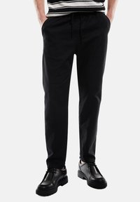 Zwarte taps toelopende broek van zachte stof, met een elastische tailleband met trekkoord en zijzakken, gecombineerd met zwarte leren schoenen.