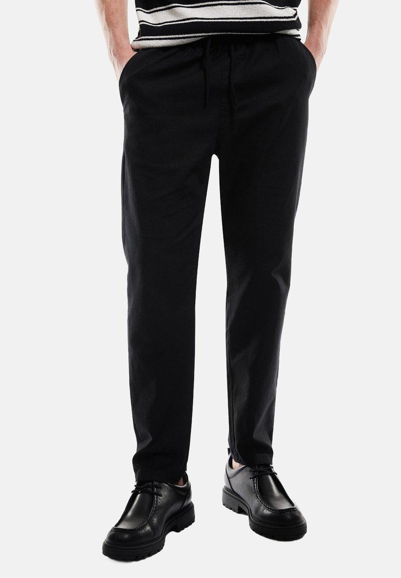Zwarte taps toelopende broek van zachte stof, met een elastische tailleband met trekkoord en zijzakken, gecombineerd met zwarte leren schoenen.