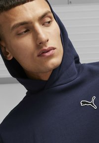 Marineblaue Kapuzenjacke aus Baumwollmischmaterial mit einer Kapuze zum Zuziehen und gesticktem Logo auf der Brust. Geschmeidige Textur, minimalistisches Design.