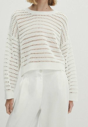 Femme portant un pull en crochet blanc à manches longues, légèrement court, associé à un pantalon blanc taille haute.