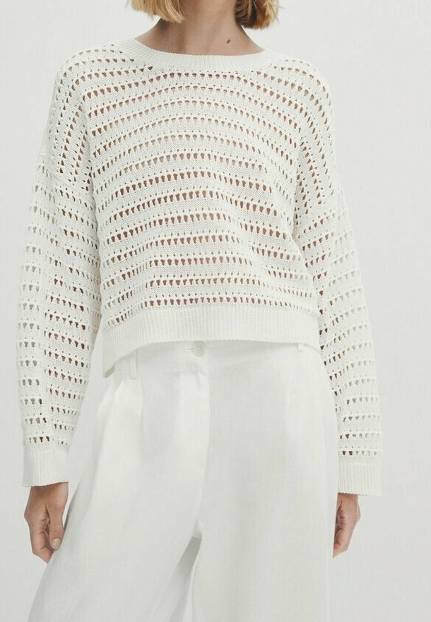Femme portant un pull en crochet blanc à manches longues, légèrement court, associé à un pantalon blanc taille haute.