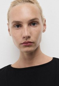 Detailní záběr na osobu se světlou pletí, rovnými blond vlasy a pihami, oblečenou v černém tričku s krátkým rukávem na bílém pozadí.