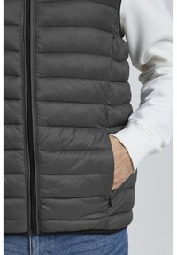 Chaleco acolchado gris con secciones de estilo acolchado horizontal, cierre de cremallera de longitud completa y bolsillos laterales. Dobladillo cortado con un acabado limpio. Interior sin características.