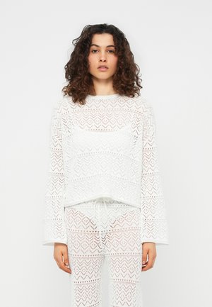 Femme aux cheveux bouclés portant un haut à manches longues blanc à motifs et un pantalon assorti, debout devant un fond blanc uni.