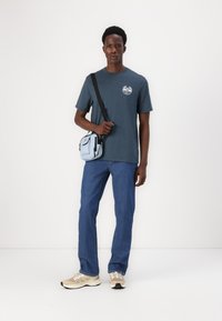 Dark teal short-sleeve t-shirt with a circular logo, paired with light blue shoulder bag, blue jeans, and beige sneakers with reflective accents.