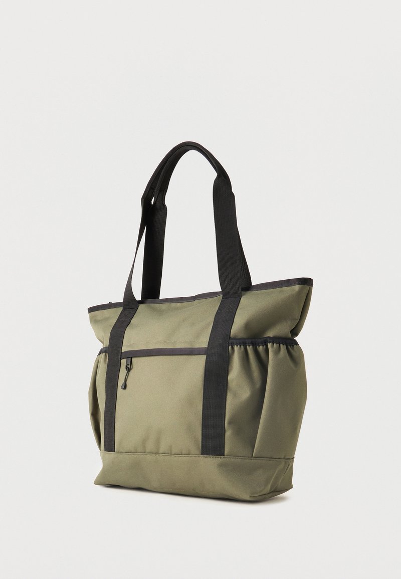 Bolsa tote verde oliva con correas negras, bolsillo frontal con cremallera y bolsillos elásticos laterales, de pie sobre fondo blanco.