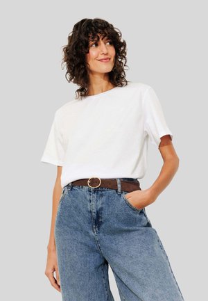 Femme aux cheveux bouclés portant un t-shirt blanc rentré dans un jeans bleu taille haute avec une ceinture marron, se tenant avec une main dans la poche.