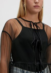 Junge Frau in einem transparenten schwarzen Langarmoberteil mit schwarzem, blickdichtem Cami und dekorativen Schleifen, kombiniert mit hellblauen Jeans.