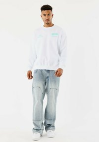 Sudadera blanca con diseños gráficos en azul claro, combinada con unos vaqueros holgados en azul claro que tienen dos bolsillos frontales y unas zapatillas blancas.