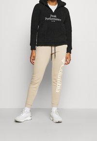 Svart fluffig hoodies med texten "Peak Performance", tillsammans med beige joggers med "Columbia"-logotyp. Vita sportskor.