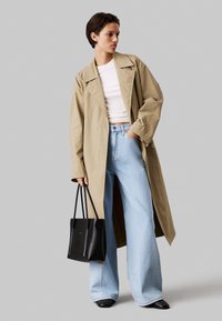 Beiger Trenchcoat mit weiten Ärmeln, kombiniert mit einem weißen, taillierten Oberteil, hellblauen weitgeschnittenen Jeans und einer schwarzen Tote-Tasche. Schwarze Schuhe sind sichtbar.