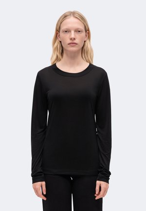 Femme aux longs cheveux blonds portant un t-shirt noir uni à manches longues, debout devant un fond gris clair.