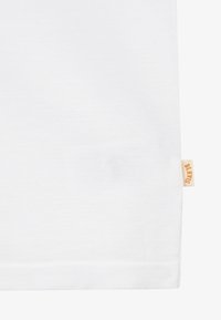 Section de tissu blanc avec une petite étiquette beige cousue sur le bord, affichant le texte orange « BLKVIS » sur l’étiquette.