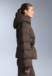 Hnedá nadýchaná bunda s kapucňou, s obtiahnutým pásom, prešívaným dizajnom a elastickými manžetami. Hladká textura a stredná dĺžka.