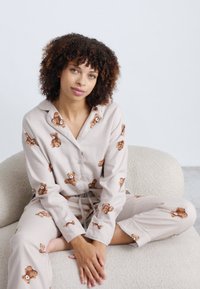 Pysjamasett i lysebeige stoff med broderte brune teddybjørner. Har en knapp-up topp og bukser med snøring, som gir en avslappet passform.