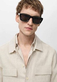 Črne pravokotne sončne očali, svetlo bež lanena srajca z dvema žepoma na prsih, z gumbi na sprednji strani, kratek ovratnik, mehak material, minimalistična estetika.