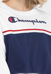 Sudadera blanca y azul marino con rayas rojas, que presenta el logo de "Champion" en azul marino. Confeccionada en un tejido suave con un corte holgado y cuello redondo.