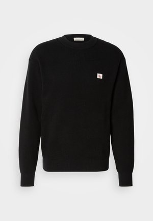 Zwarte gebreide trui met ribbeltextuur, ronde hals, en een klein wit en rood logo-opdruk op de linkerkant van de borst. Classieke pasvorm met lange mouwen.