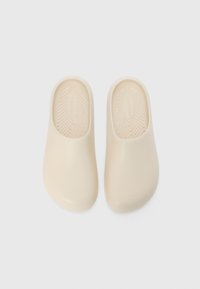 Beige slip-on clogs made of smooth material, featuring a textured insole with small dots. Rounded toe and contoured heel for comfort.