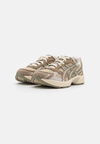 Beige og tan atletiske sko med mesh-paneler, kremfargede snørebånd og teksturerte gummisåler; har en subtil logo og støttende design.