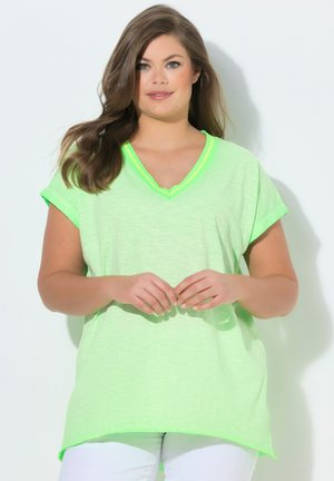 Frau mit langen braunen Haaren, die ein lockeres neongrünes V-Ausschnitt-Shirt und weiße Hose trägt, steht mit sanft verschränkten Händen.