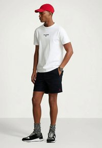 T-shirt branca com o logótipo "Polo Sport", calções azul-marinho com logótipo, boné vermelho, meias com padrão e sapatilhas pretas com detalhes brancos.