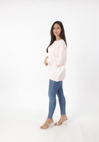 Lichtroze gebreide trui met ballonmouwen, gecombineerd met blauwe skinny jeans en doorzichtige blokhak-sandalen. Geplaatst tegen een witte achtergrond.