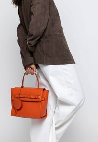 Orangefarbene Lederhandtasche mit strukturiertem Design, oberen Griffen und einem abnehmbaren runden Accessoire. Getragen mit einer braunen Wildlederjacke und weißen Hosen.