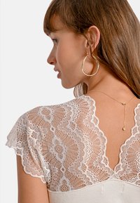 Top bianco in pizzo con motivi floreali e bordi scallop; presenta un corpo in tessuto morbido e dettagli delicati sul retro. Collana in catena dorata visibile.