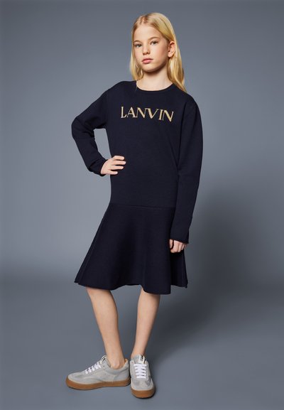 Ναυτική πλεκτή φόρεμα με εφαρμοστό σώμα, φουσκωτή φούστα και λογότυπο "LANVIN" σε χρυσό. Γκρίζα αθλητικά παπούτσια με λευκές κορδόνες και υφασμάτινη σόλα.
