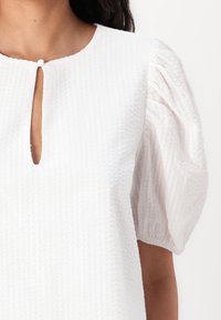 Blouse blanche texturée avec un col rond et une petite ouverture en forme de clé, dotée de manches bouffantes et d'un tissu à motif subtil.