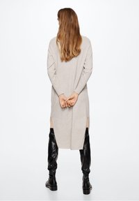Femme aux longs cheveux châtain clair portant une robe beige à manches longues avec des fentes latérales et des bottes noires montantes jusqu’aux genoux, debout les mains croisées dans le dos.