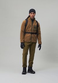 Brązowa polarowa kurtka z zieloną kieszenią, czarne rękawice, oliwkowe spodnie i czarne buty. Model nosi czarny plecak i czapkę beanie w kolorze czarnym.