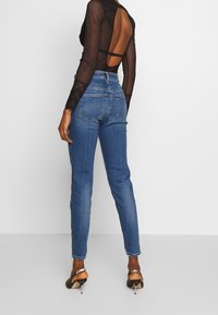 Džíny s vysokým pasem modré barvy, úzkého střihu, s pěti kapsami a lehkým vyblednutím podél nohavic, spárované s černým průsvitným topem s dlouhým rukávem.