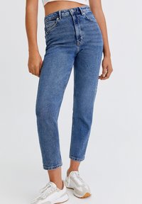Jeans de tiro alto en azul con un acabado desgastado, cinco bolsillos y un corte recto, combinados con zapatillas blancas.