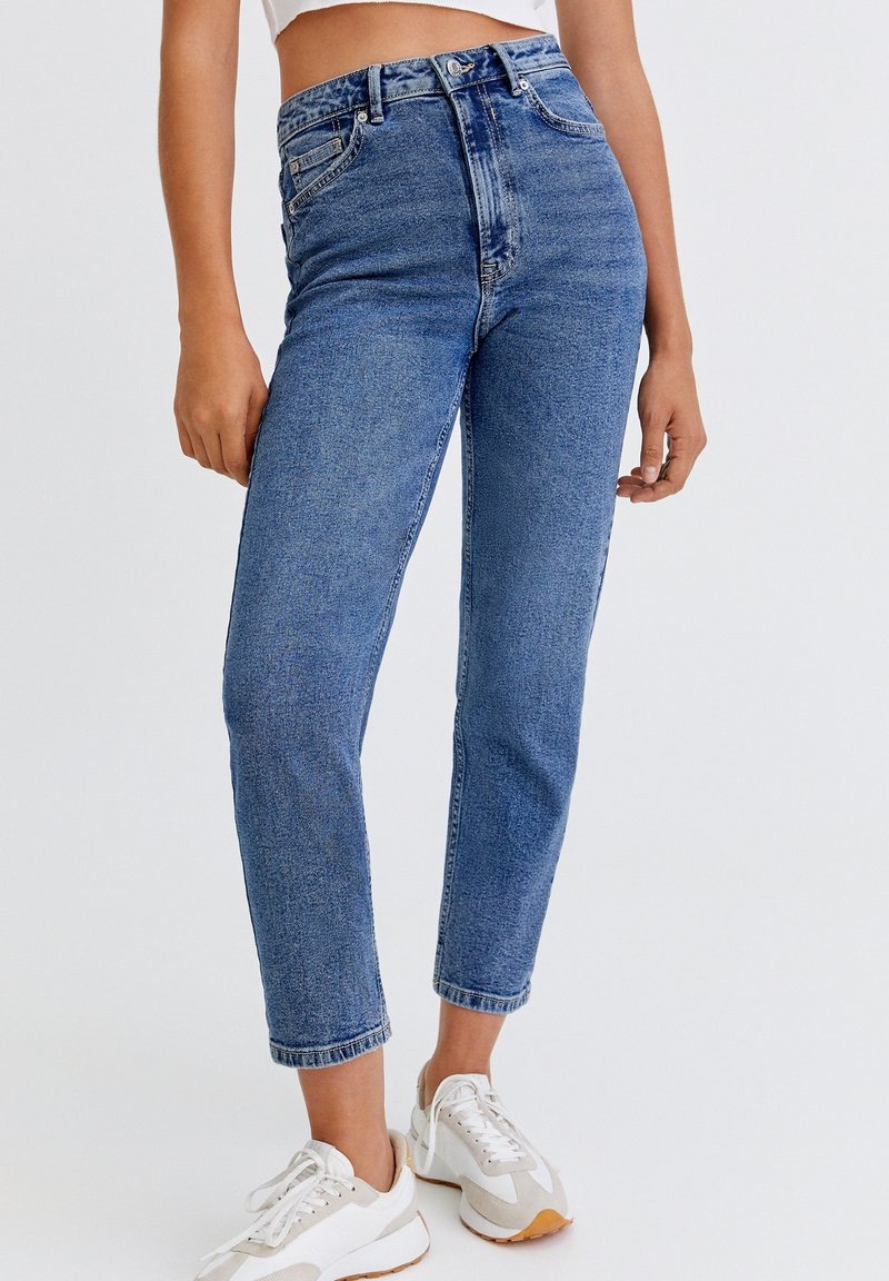 Jeans de tiro alto en azul con un acabado desgastado, cinco bolsillos y un corte recto, combinados con zapatillas blancas.