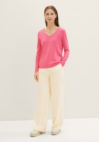 Pull élégant rose vif à col en V avec des manches longues, associé à un pantalon large jaune pâle. Le mannequin se tient devant un fond clair.