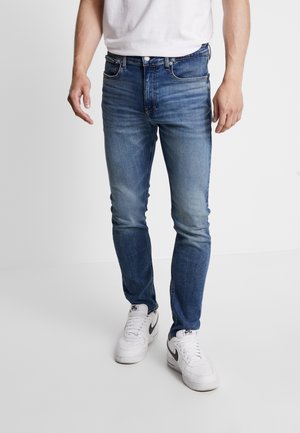Mann trägt ausgewaschene blaue Slim-Fit-Jeans und weiße Nike-Sneaker, steht vor einem schlichten hellen Hintergrund.
