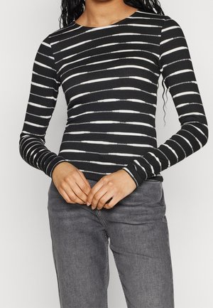 Top nero a maniche lunghe con righe orizzontali bianche, texture a coste, scollatura tonda e design aderente, abbinato a jeans grigi.