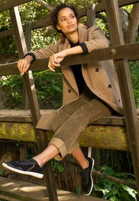 Brown checked coat with black buttons, paired with olive green pants and black sneakers. The setting features wooden structures and greenery.
