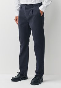 Pantalon bleu marine en tissu léger, avec une coupe droite, des plis à l'avant et des poches latérales. Associé à des chaussures noires.