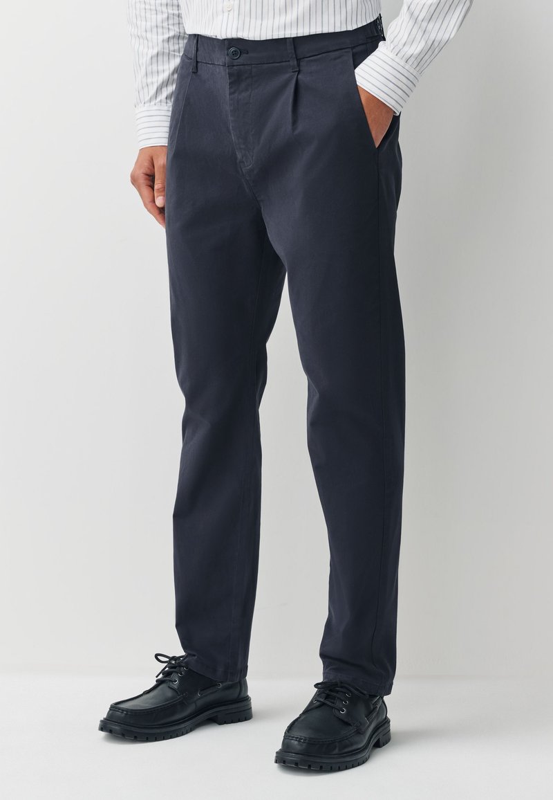 Pantalon bleu marine en tissu léger, avec une coupe droite, des plis à l'avant et des poches latérales. Associé à des chaussures noires.
