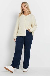 Krämfärgad stickad cardigan med rund halsringning, knäppning med knappar och två framfickor, ihop med mörkblå utsvängda jeans och vita sneakers.
