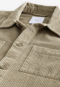 Chemise en velours côtelé beige avec col à rabat, deux poches avant et boutons contrastants. Tissu texturé avec nervures verticales.