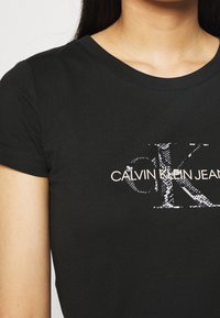 Czarna bawełniana koszulka z okrągłym dekoltem; ma biały logo "CALVIN KLEIN JEANS", z teksturowanym wzorem węża na literze "K". Krótkie rękawy.