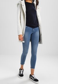 Abrigo gris con capucha, camiseta azul marino, jeans skinny azul claro, zapatillas negras y blancas. Texturas suaves, diseño casual, sin patrones ni herrajes visibles.