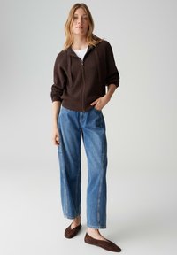 Hoodie à fermeture éclair côtelée marron, t-shirt blanc, pantalon en denim bleu ample et ballerines marron. Le mannequin est debout, une main dans la poche, fond neutre.