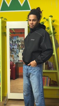 Czarna kurtka na zamek z szarymi akcentami i wysokim kołnierzem, w połączeniu z jasnoniebieskimi dżinsami; stoi przed lustrem na żółtej ścianie.