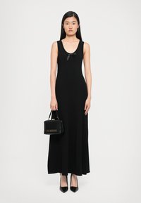Woman in a sleeveless black maxi dress with a small bow at the neckline, holding a black Love Moschino handbag and wearing black heels.